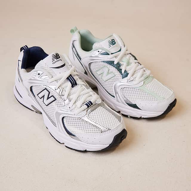 A pair of white trainers with mesh detailing and distinct branding on the side. The shoes have chunky soles and green accents, with one shoe lined in green and the other in navy. They are positioned on a light-coloured surface.