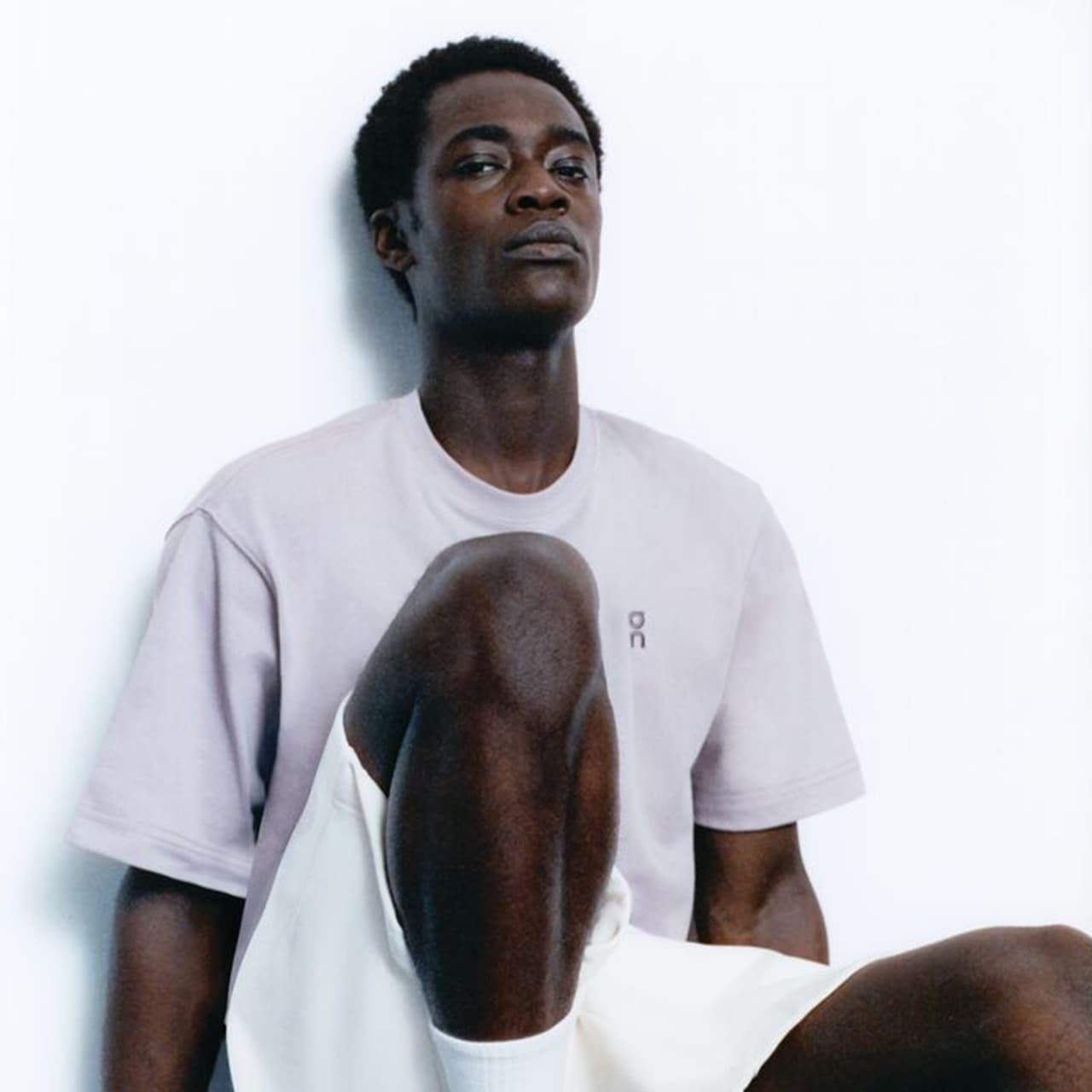 A person sitting with one knee raised, wearing a light purple T-shirt and white shorts. Their expression is neutral, and they are positioned against a plain background, creating a minimalist and relaxed atmosphere.