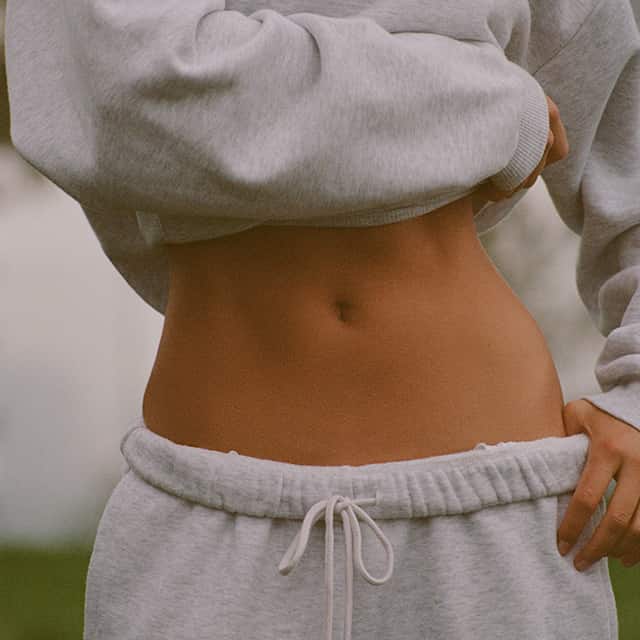 A person wearing a grey cropped sweatshirt and grey drawstring tracksuit bottoms, showing their midriff. The background is blurred.