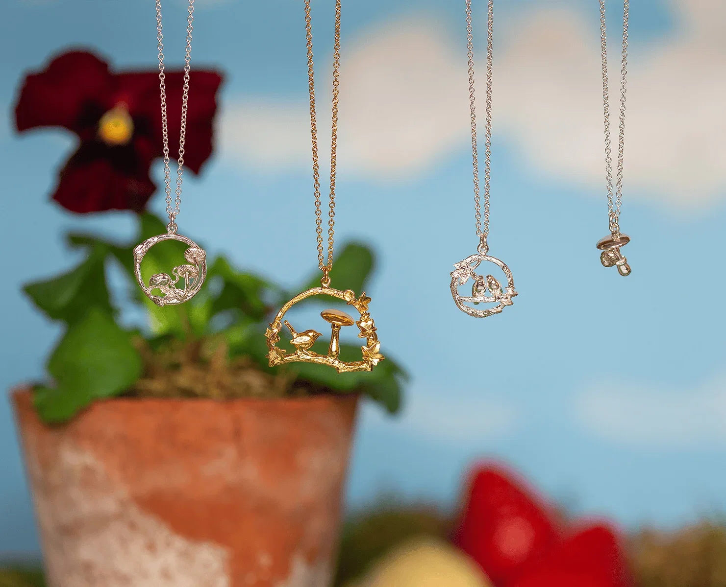 Delicate nature-inspired necklaces hang against a softly blurred garden backdrop. The jewelry features intricate woodland scenes, including mushrooms, birds, and small animals, crafted in silver and gold tones. A terracotta flower pot and deep red flowers appear out of focus in the background, creating a whimsical, earthy atmosphere.
