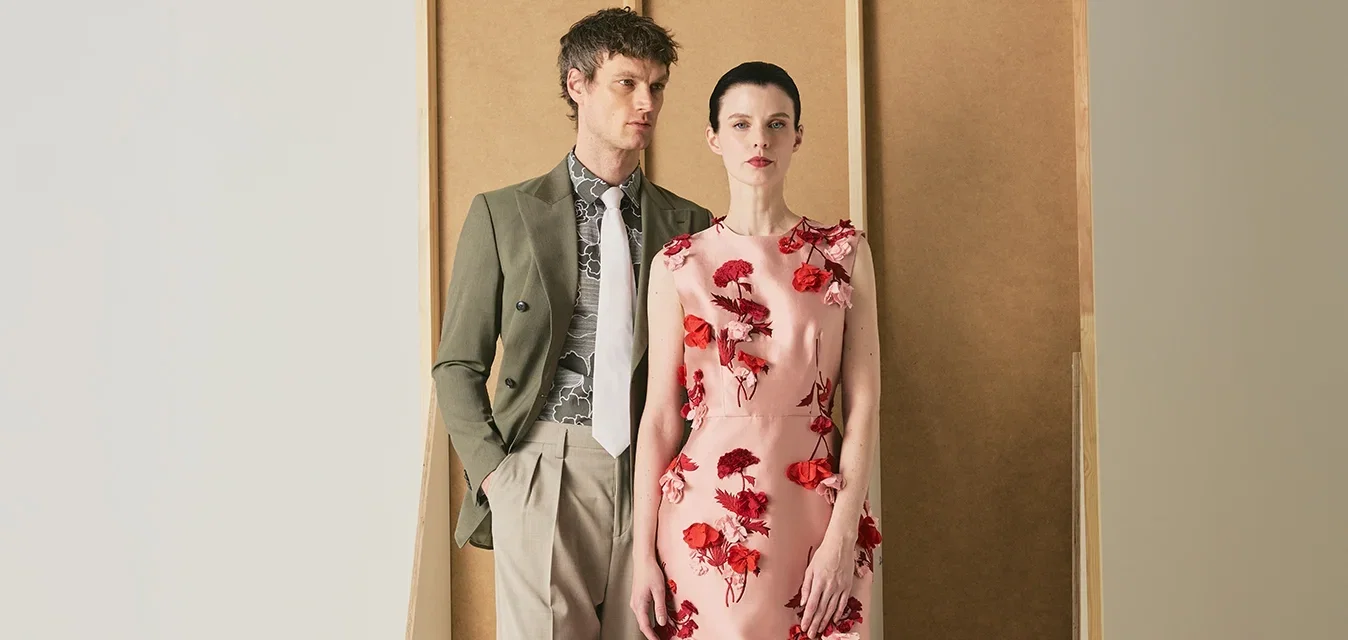 Ein Mann in olivgrünem Blazer und beiger Hose steht neben einer Frau in einem rosa Blumenkleid vor einem minimalistischen beigen Hintergrund.