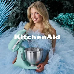 Vrouw in een volumineuze lichtblauwe tulejurk die een muntgroene standmixer vasthoudt, omringd door blauwgroene bladeren en cupcakes op een klein tafeltje.