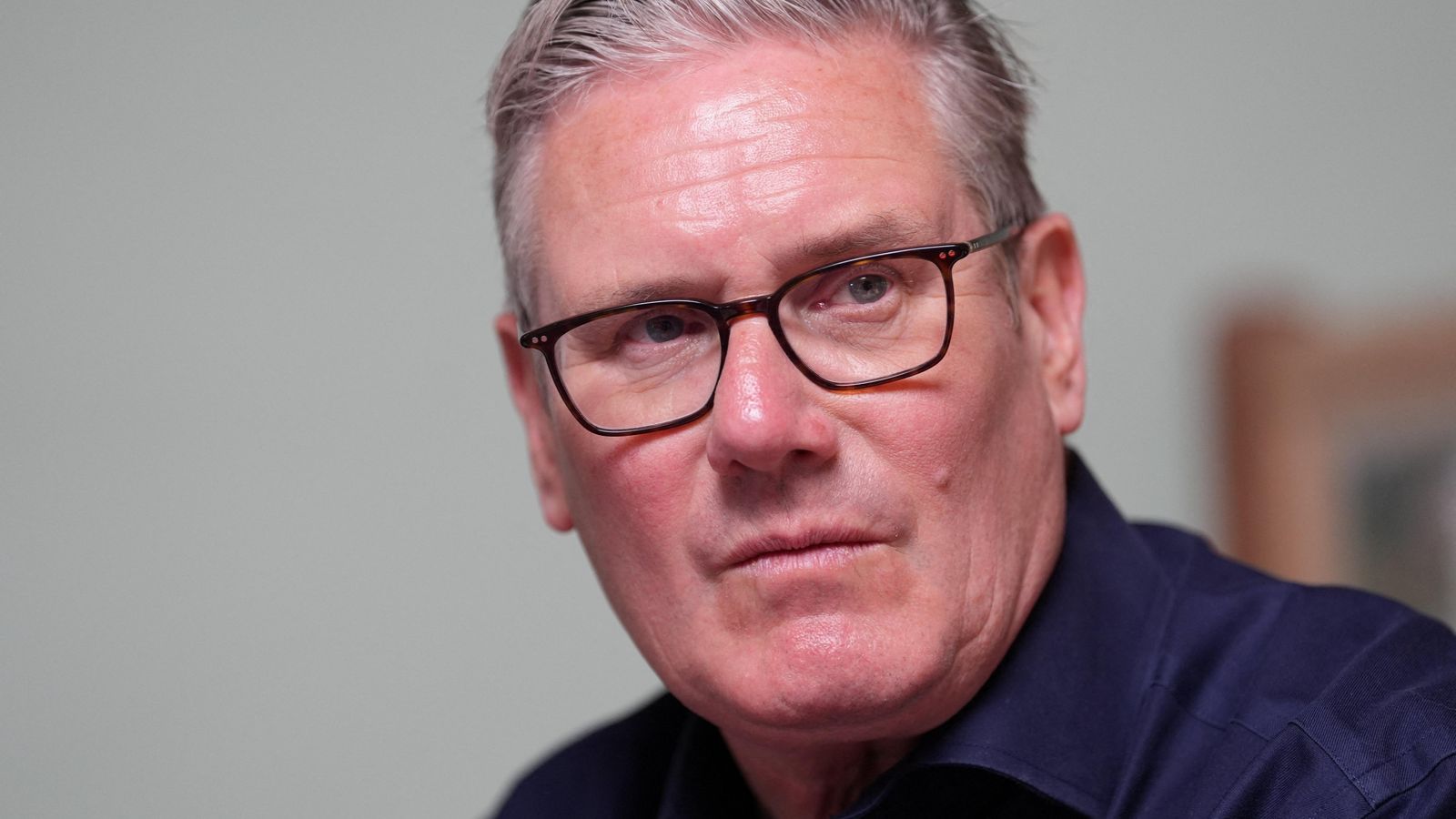 Sir Keir Starmer wearing glasses and a dark blue shirt. He looks sternly to the right of the camera and has grey hair.