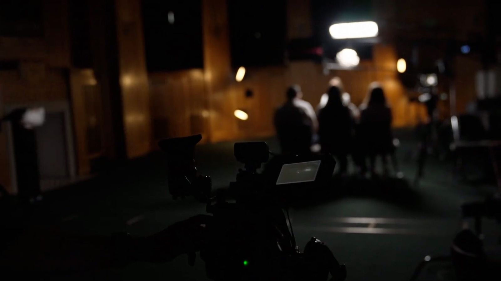 A darkened room with several figures in the background, out of focus.