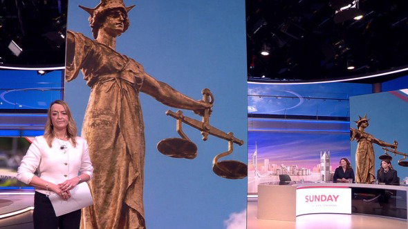 Laura Kuenssberg stands in a studio wearing a white top and dark trousers. On the screen behind her is a picture showing the scales of justice; interviewee Darren Jones is on her right and the panel are to her left.