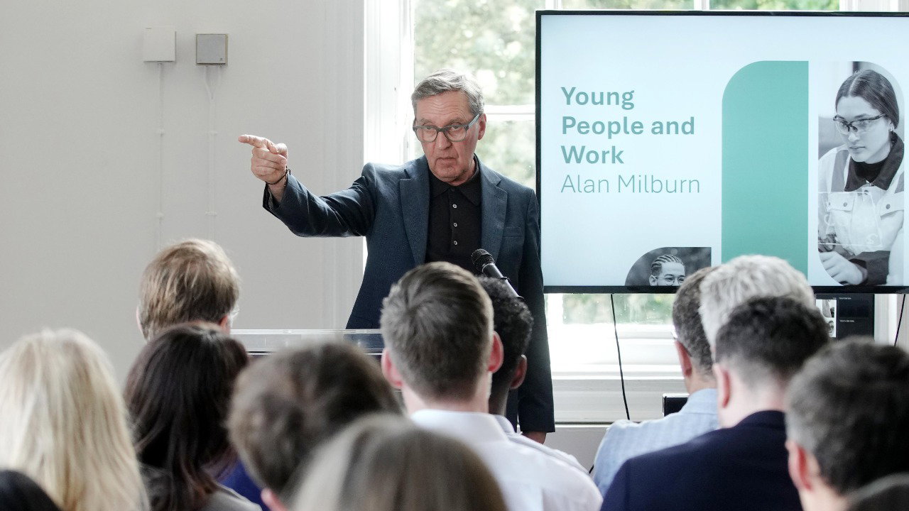 Former health secretary Alan Milburn speaks to the media on the publication of the interim Milburn Report into Young People and Work, at West Library Youth Employment Hub, north London. He stands pointing with his index finger, wearing glasses, a dark shirt and blue jacket. Behind him, text reads: ‘Young People and Work Alan Milburn’.