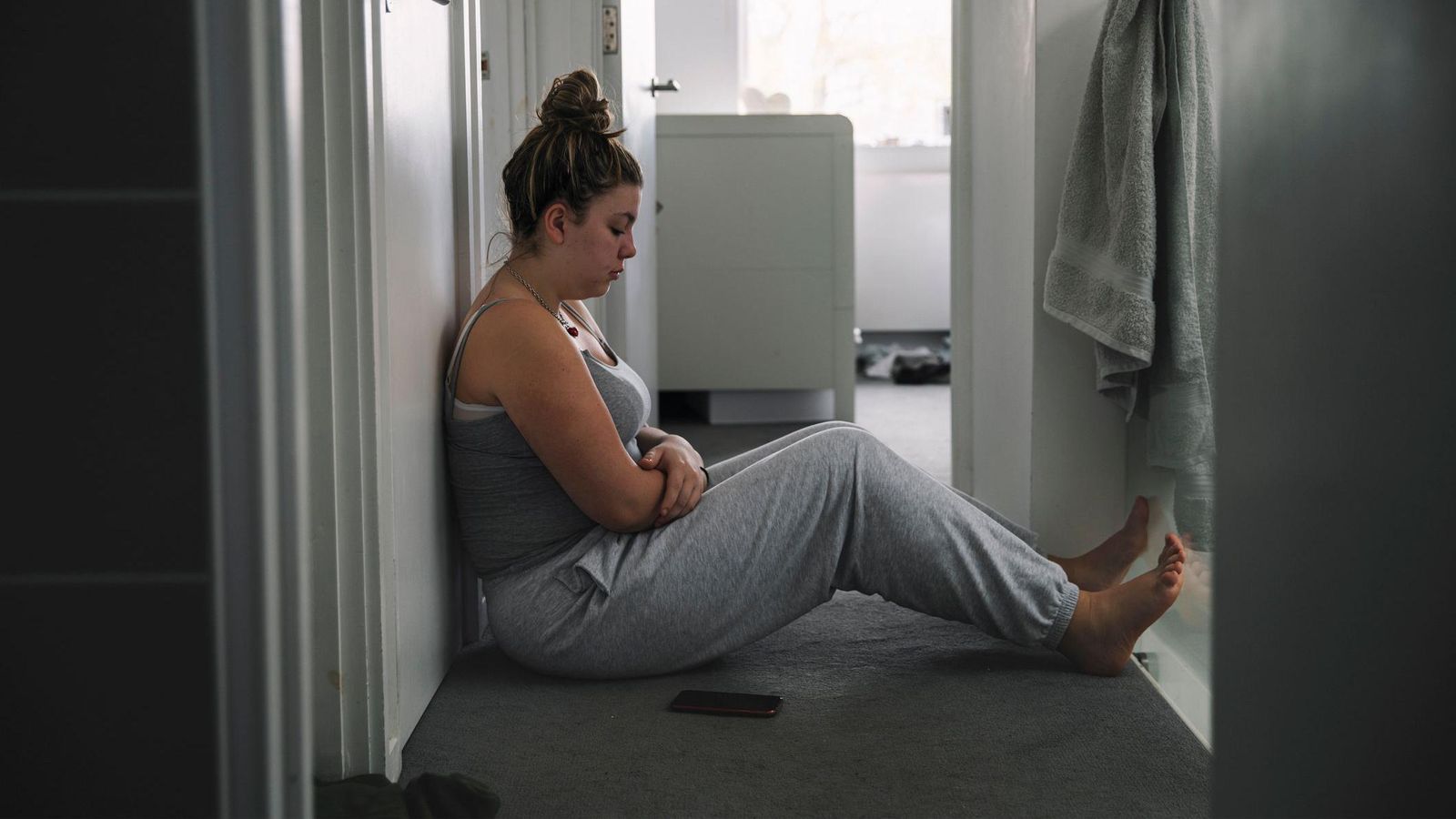 A young woman sits in the hallway of a home looking sad and lonely, with her phone on the floor beside her. She wears grey tracksuit bottoms and a grey sleeveless top and has her hair in a bun.