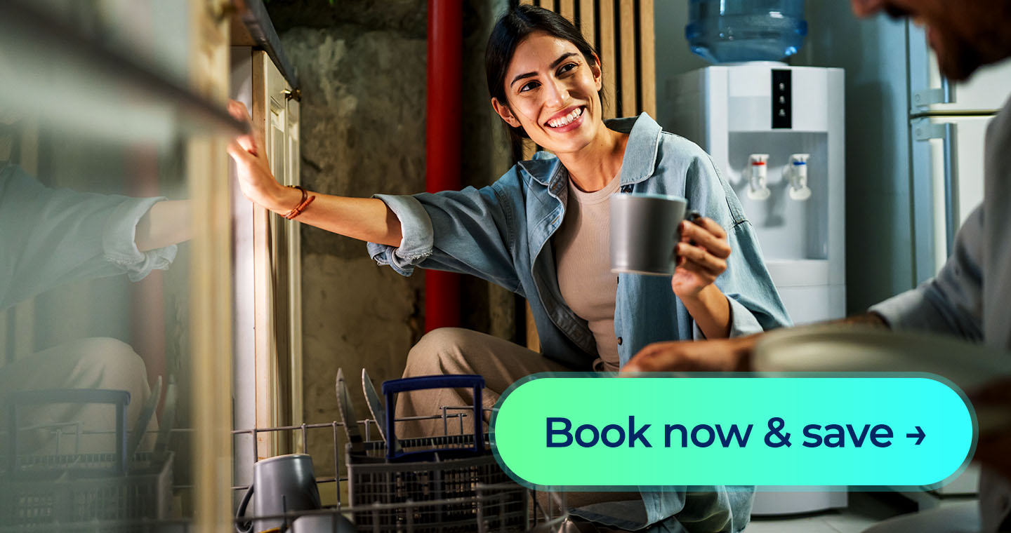 A woman smiling while loading cups into a dishwasher in her kitchen, with a 'Book now & save' button displayed in front