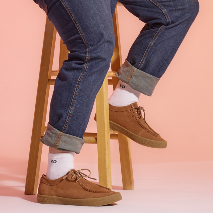 Man's legs from the waist down sitting on a stool wearing jeans, white socks with the Wide Open logo and brown shoes with a peach background.