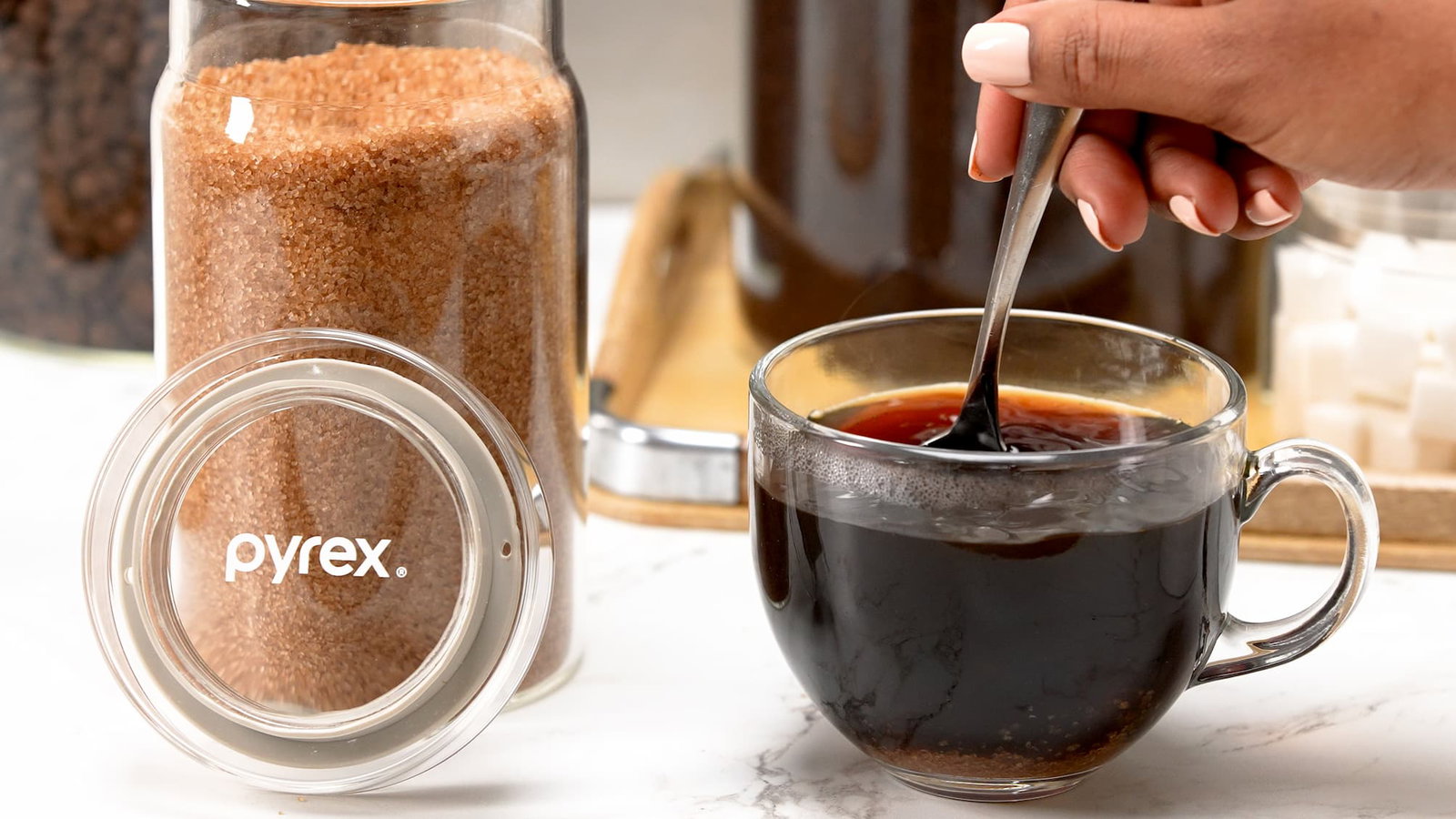 Glass canister with brown sugar next to a cup of coffee being stirred.