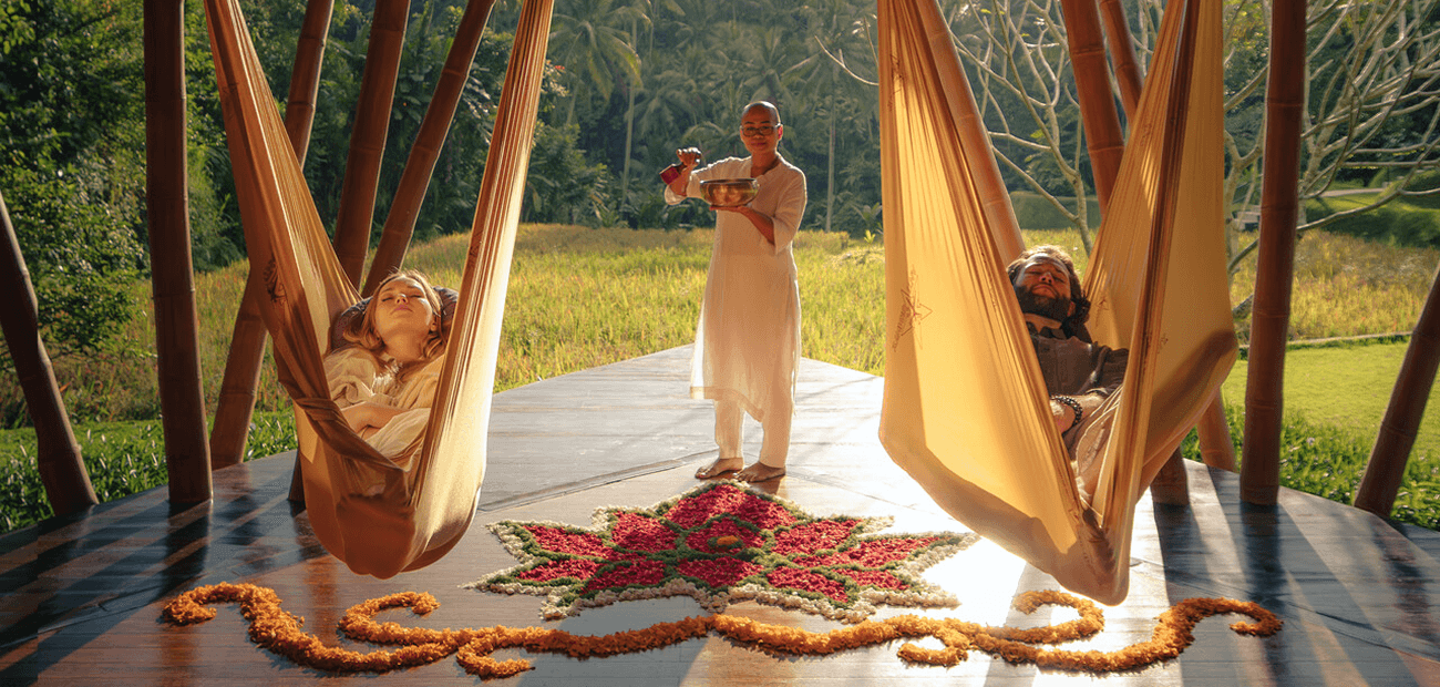 Two people relax in hammocks while a staff member with a sound bowl stands nearby.