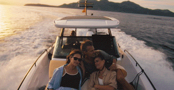 Hero video featuring 5 scenes: 1) Family enjoying a boat ride at sunset with mountains in the background. 2) Hot air balloons drifting above fog-covered pyramids. 3) Woman receiving a ceremonial blessing from a man holding a clay pot with incense. 4) Hands gently pass over a person resting in a hammock. 5) Staff member serving plates of food at an outdoor dining and cooking area.