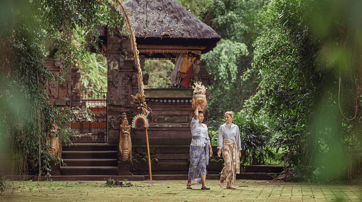 Two women walk near a temple, one carrying a decorative offering.