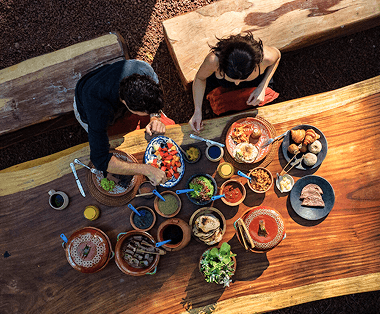Two people enjoy food at a large wooden table outdoors.