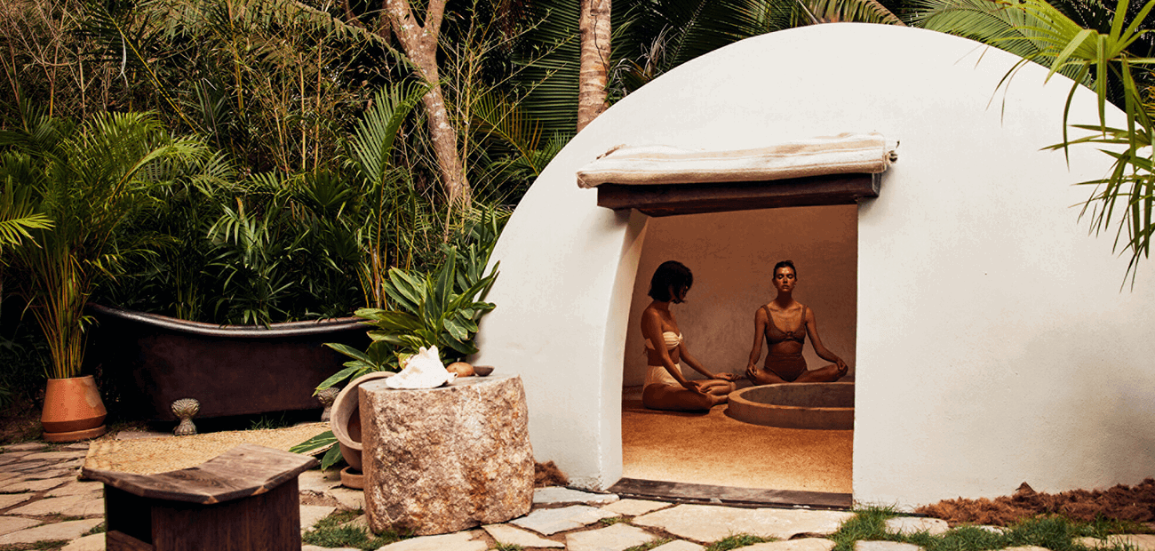 Two women meditating in a round, white structure in a lush garden.