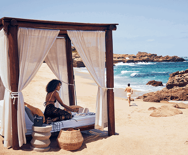 Woman sitting on a beach cabana with a man walking along the shore.