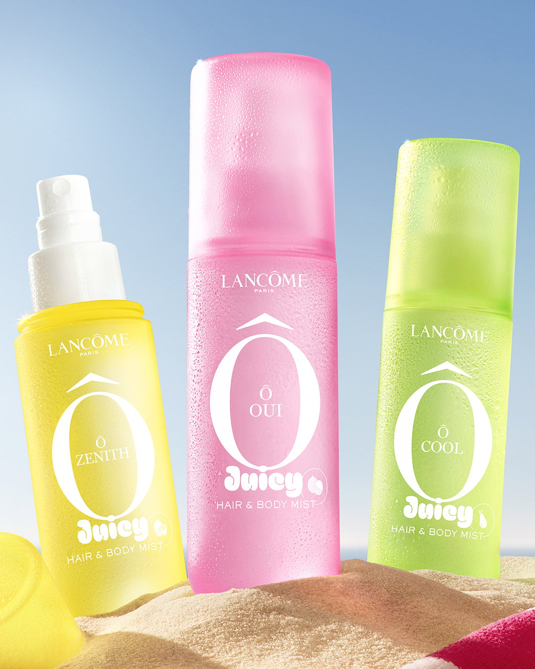 Le tre nuove Hair & Body Mists Lancôme, Ô Zenith, Ô Oui e Ô Cool, su una spiaggia sabbiosa con cielo azzurro sullo sfondo.