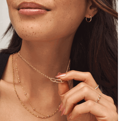 Woman wearing layered gold chain necklaces, small hoop earring, and delicate rings, holding necklace clasp.