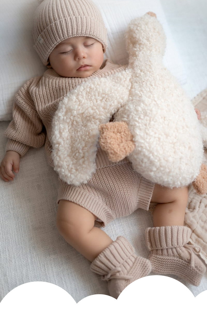 Sleeping baby wearing a knit hat and beige outfit while holding a large cream plush duck toy on a soft blanket.