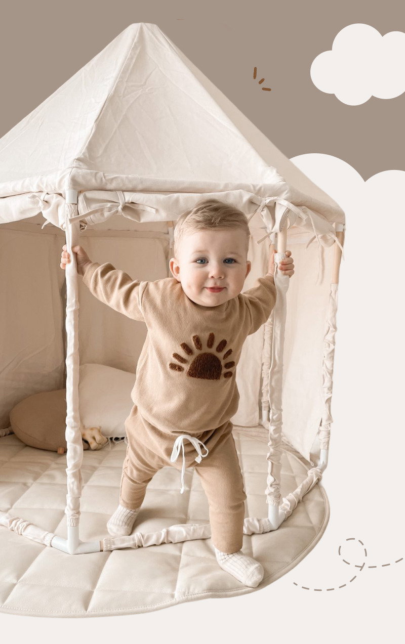 Smiling toddler standing inside a soft cream play tent while wearing a matching beige lounge set with sun design details.