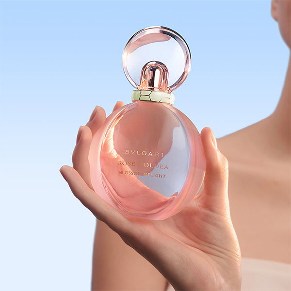 A hand holding a pink oval perfume bottle with a looped cap against a gradient blue background.