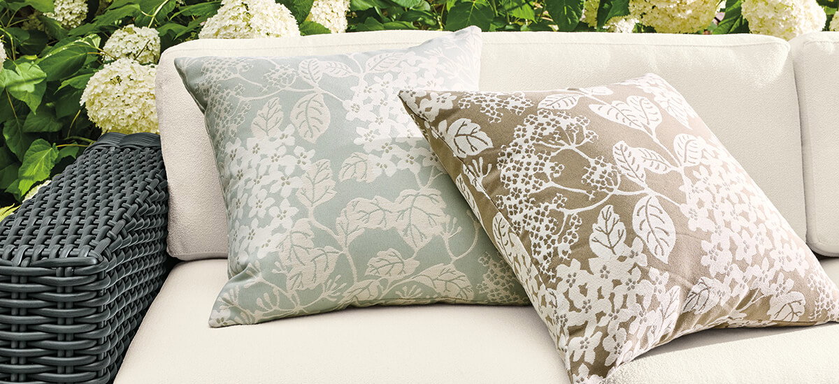Pair of patterned outdoor pillows in muted sage and taupe on a cream cushioned bench with black woven armrest.