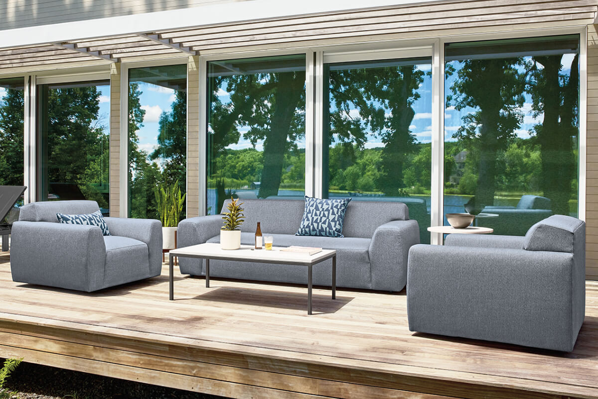 Drift outdoor sofa and lounge chairs with grey upholstery on a wood deck beside large windows.