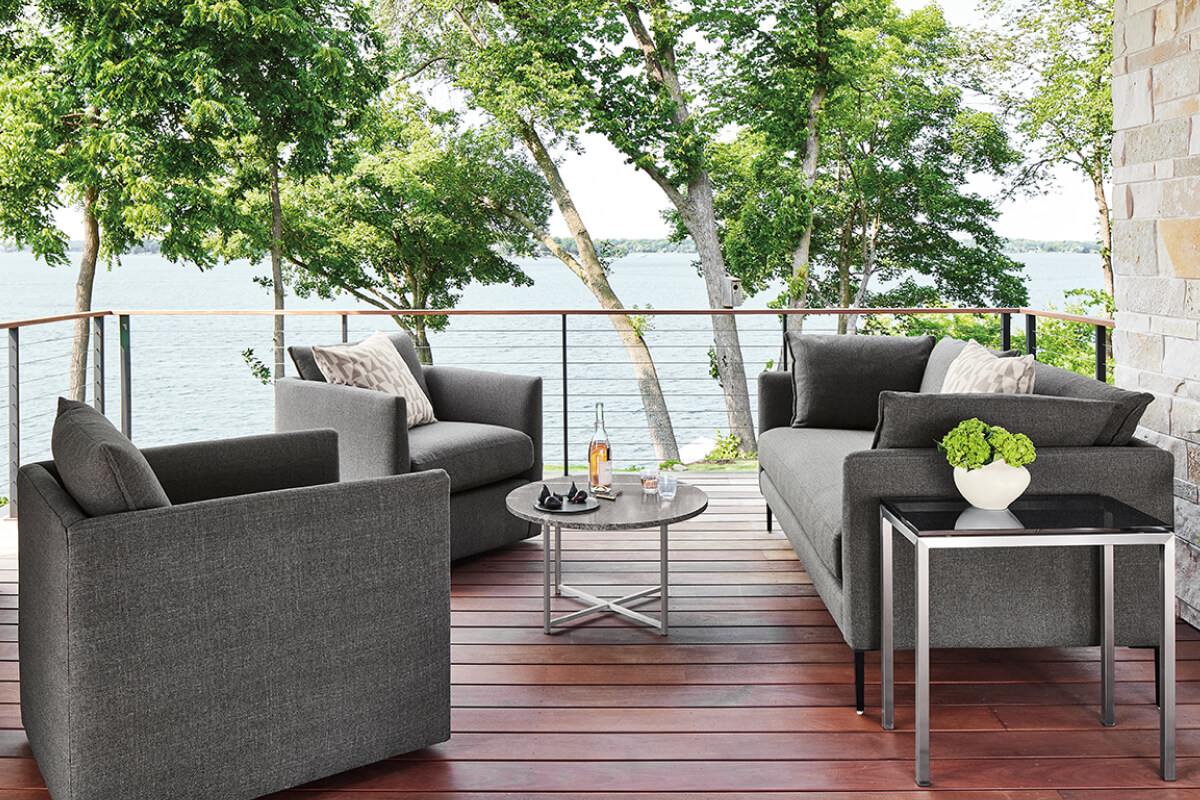 Palm sofa and lounge chairs with grey upholstery and metal tables on a lakeside deck.