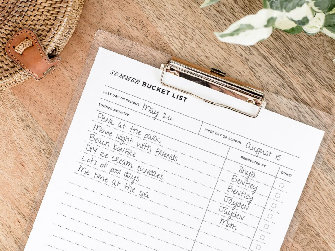 free summer bucket list printable, shown with writing, on clipboard on wood table, plant, wicker basket