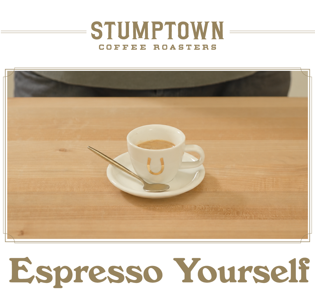 A small white espresso cup with a gold horseshoe logo sits on a saucer with a spoon on a light wood table. Text below reads “Espresso Yourself.”