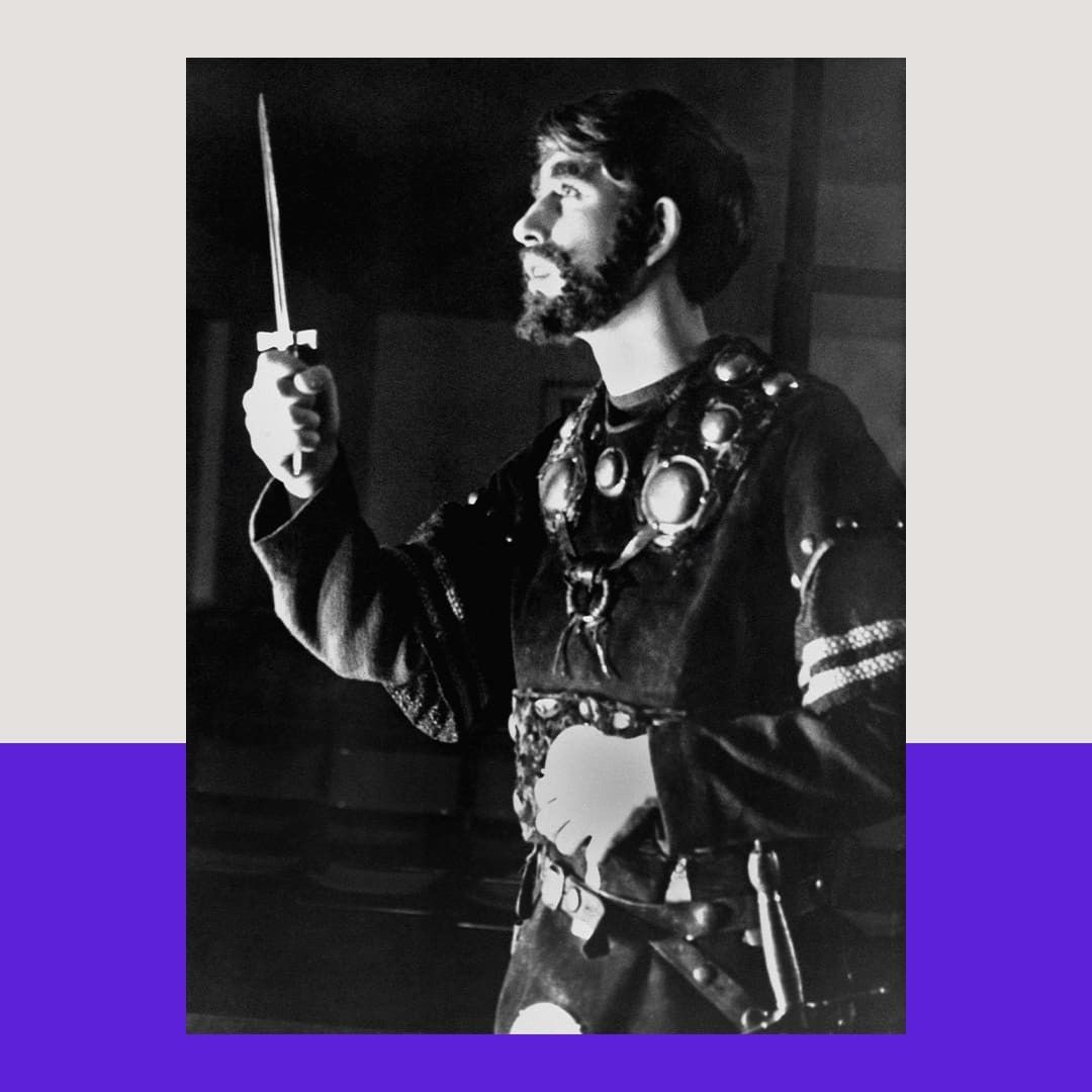 A black and white photo shows a young then-Prince Charles wearing a costume medieval army robe with a belt and a heavy necklace. One hand is on his stomach and the other lifts a prop dagger, as he looks out to the audience. He stands in profile and wears a full beard. The photo has a light beige and purple graphic border added around it.