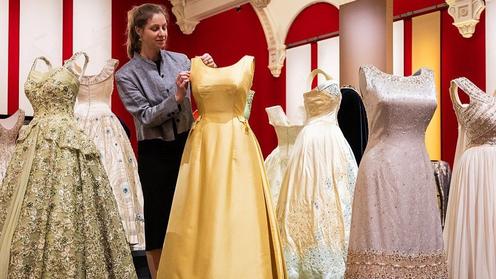 A selection of Queen Elizabeth II’s ballgowns on display at the King’s Gallery in Buckingham Palace. A member of the curation team is adjusting the strap on a yellow dress. Others include a silver dress covered in glittering beads, and a lacey green gown with a large skirt.