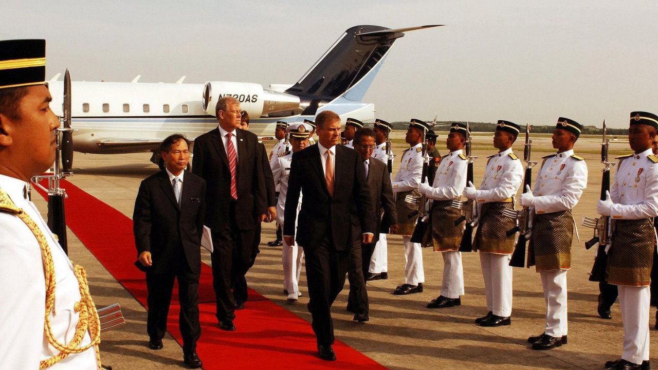 Andrew Mountbatten-Windsor walks along a red carpet on an airport tarmac, with other officials. All the men wear black suits, with a row of men dressed in white military uniform with rifles flanking the red carpet on both sides.