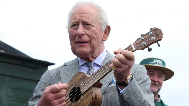 King Charles holds a wooden ukulele to his chest and grips the neck while appearing to strum. He is pictured outside on a cloudy day, wearing a grey suit with a purple shirt.