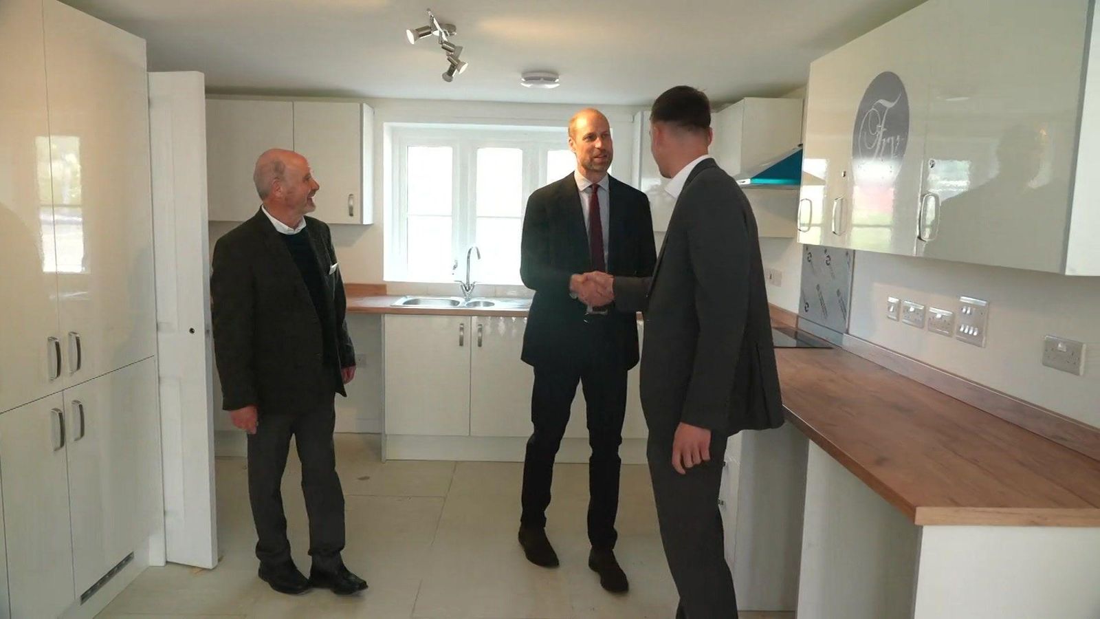 Prince William is shaking hands with a man who is standing in a newly built kitchen. The doors and cupboards are white, and the worktops are a light brown wood. There is also a man standing to the left of Prince William, smiling at him. They are all wearing black suits.