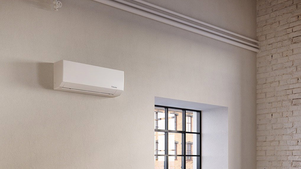 Wall-mounted white air conditioner on a pale plaster wall in a loft-style room with industrial window and exposed brick