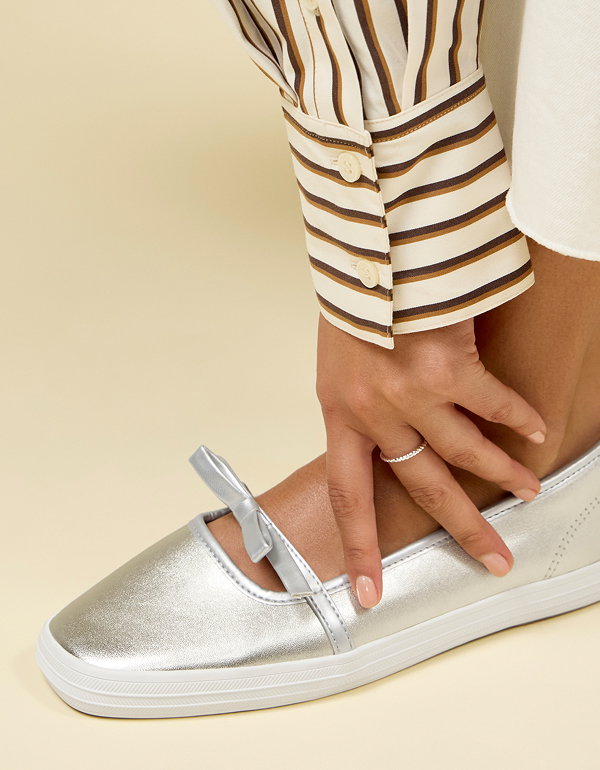 A close-up of the Square Toe Mary Jane Leather Mini Bow in champagne/silver shown on foot paired with white pants and a striped button-up.