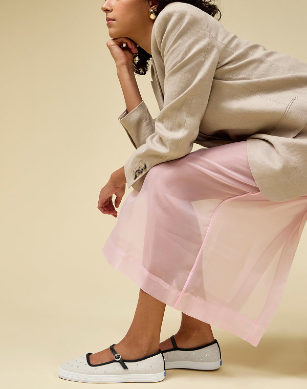 A beige background and a woman in a seated position wearing a tan blazer, sheer pink skirt, and the exclusive Square Toe Mary Jane Woven in neutral/black shoes.