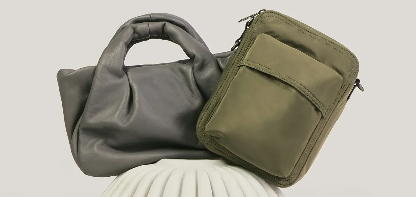 Dos bolsos, uno tote de cuero suave gris y otro bolso bandolera rectangular verde oliva, exhibidos sobre un soporte blanco con textura.
