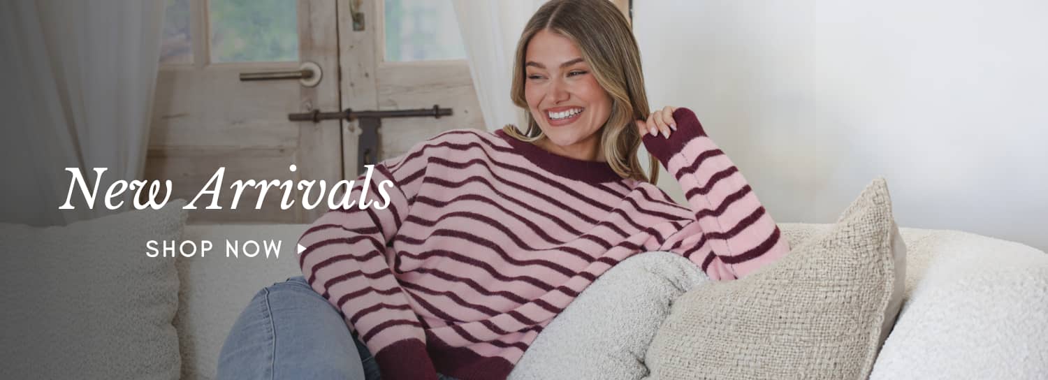 A medium shot shows a woman with blonde hair wearing a pink and maroon striped sweater and blue jeans, sitting on a couch with textured pillows, with ...