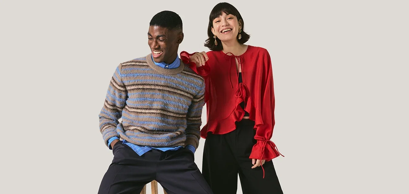 Homme en pull à rayures assis sur un tabouret, femme en blouse rouge debout à côté de lui, souriante et pointant devant elle.