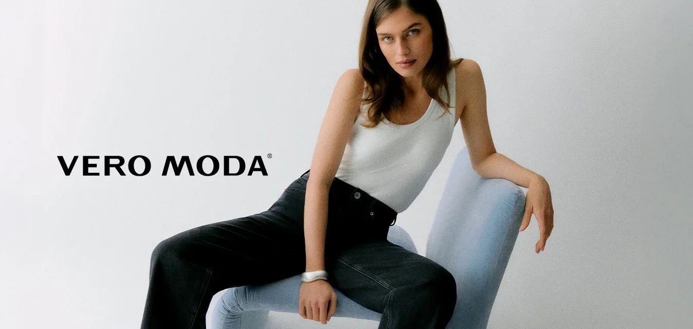 Femme aux longs cheveux bruns assise sur une chaise moderne bleu clair, portant un débardeur blanc et un jean noir à jambes larges, sur un fond blanc uni.