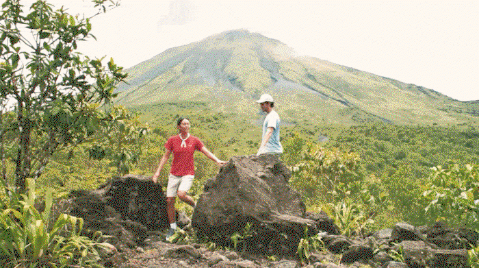 A gif of a man and woman hiking through a rainforest with a mountain in the background.