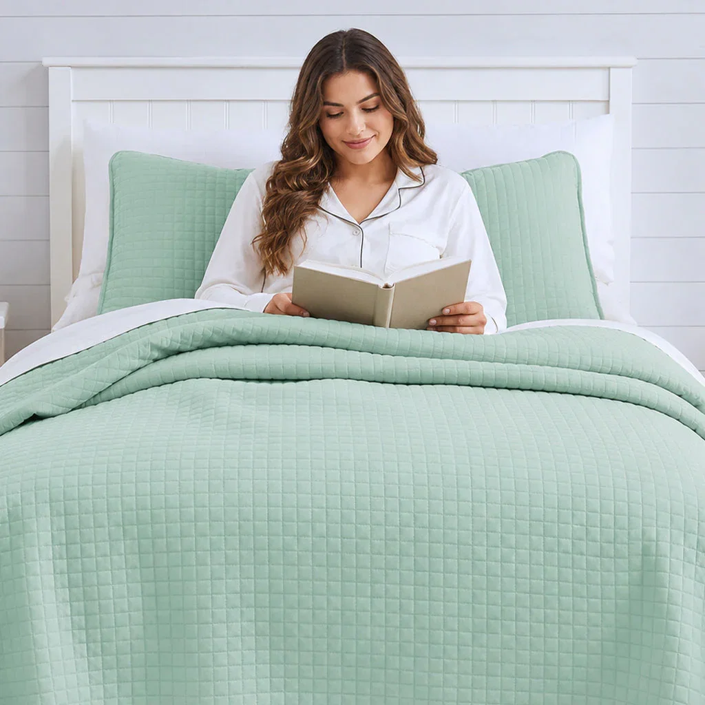 Woman reading in bed with sage green quilted bedding and white pillows in the Bedding Essentials collection.