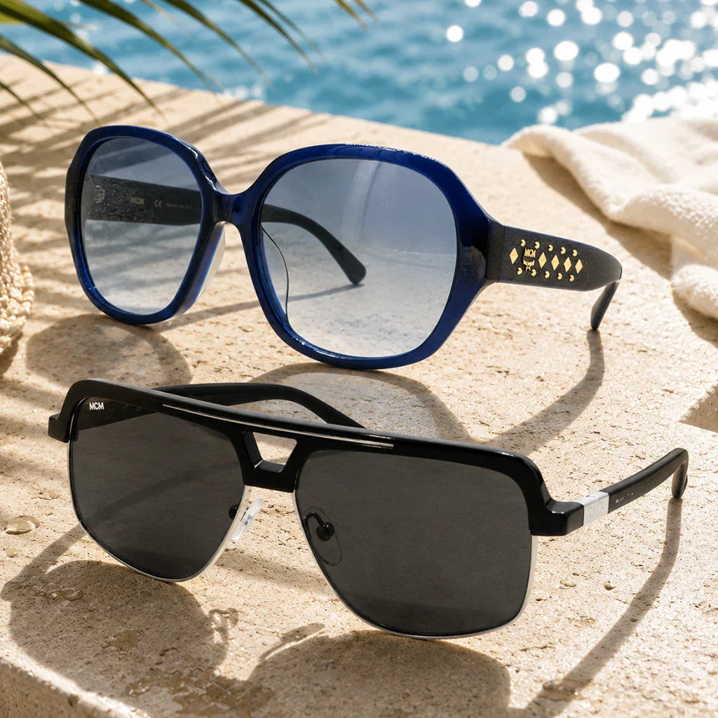 Two MCM sunglasses displayed on sand by a pool: blue oversized frames with studded temples and black aviator-style frames.