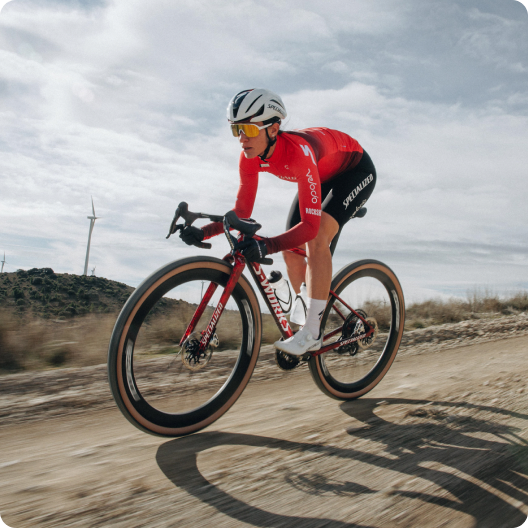 Cyclist in red kit on a red gravel bike riding a dusty country road past wind turbines under a cloudy sky