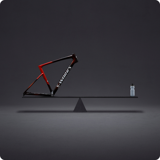 Red and black S-Works carbon road bike frame balanced on a seesaw beam opposite a water bottle in a minimal grey studio