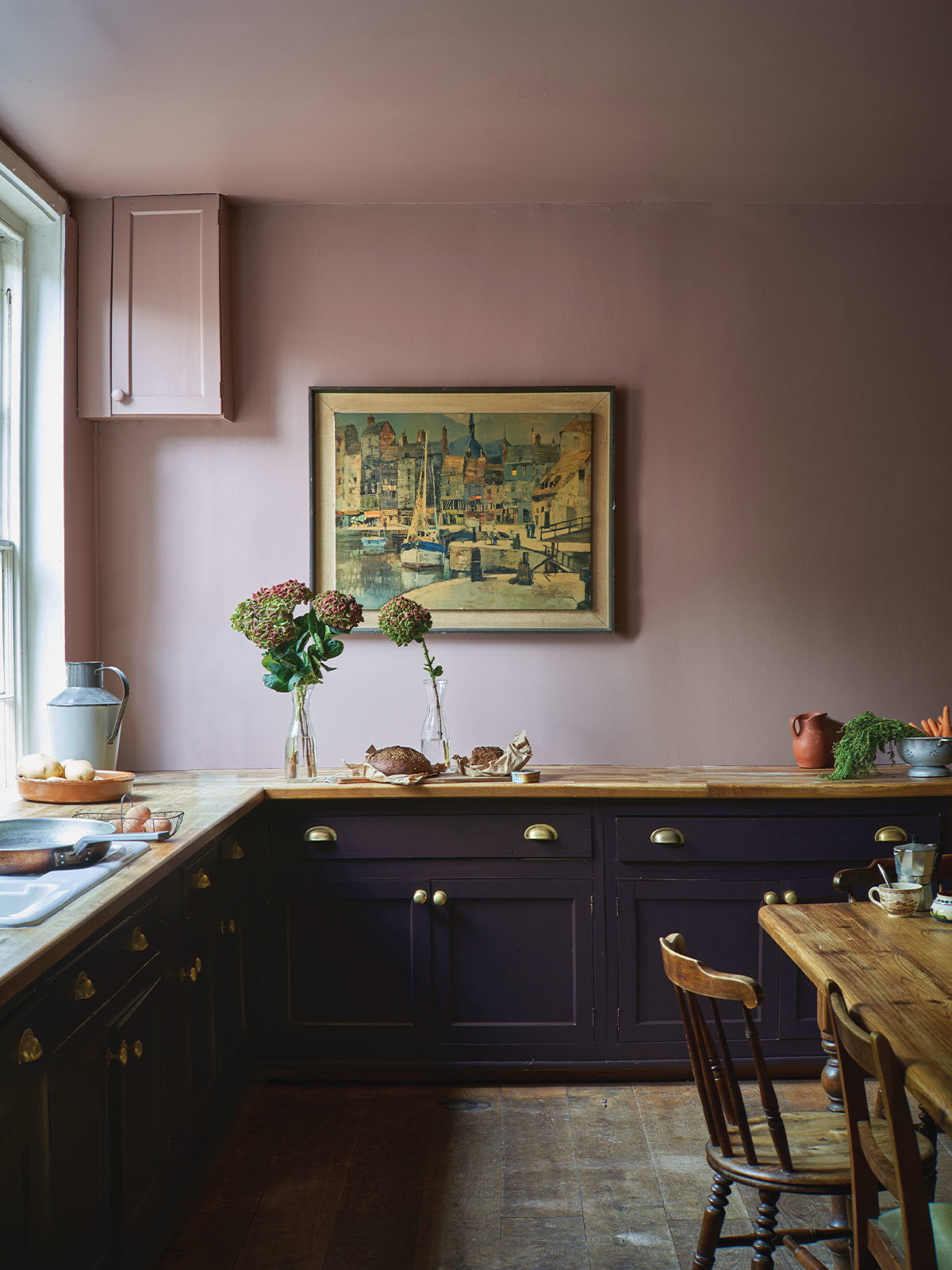 That pause in a kitchen just before you start preparing lunch. Gentle light from the window lights up the pale mauve coloured walls and wooden worksurfaces. There’s sourdough bread waiting to be sliced, eggs ready to be cooked, and a wooden table ready to linger around. The brass handles reflect gently against the dark coloured painted cabinets.