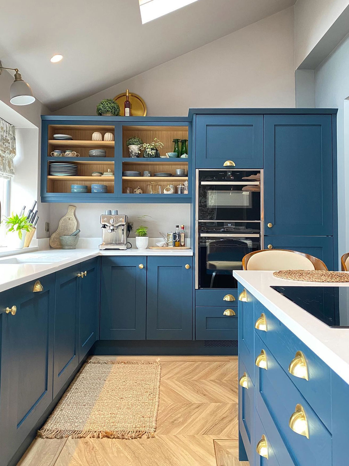 The impact of this kitchen comes from its vivid blue painted cabinets, offset with the gentle gleam of brass handles and doorknobs. The gleaming white worksurfaces provide contrast to the blue, with hints of green from the window blind, houseplants and glassware. The natural light from the window and skylight stop the rich colour from making the room feel smaller or darker.