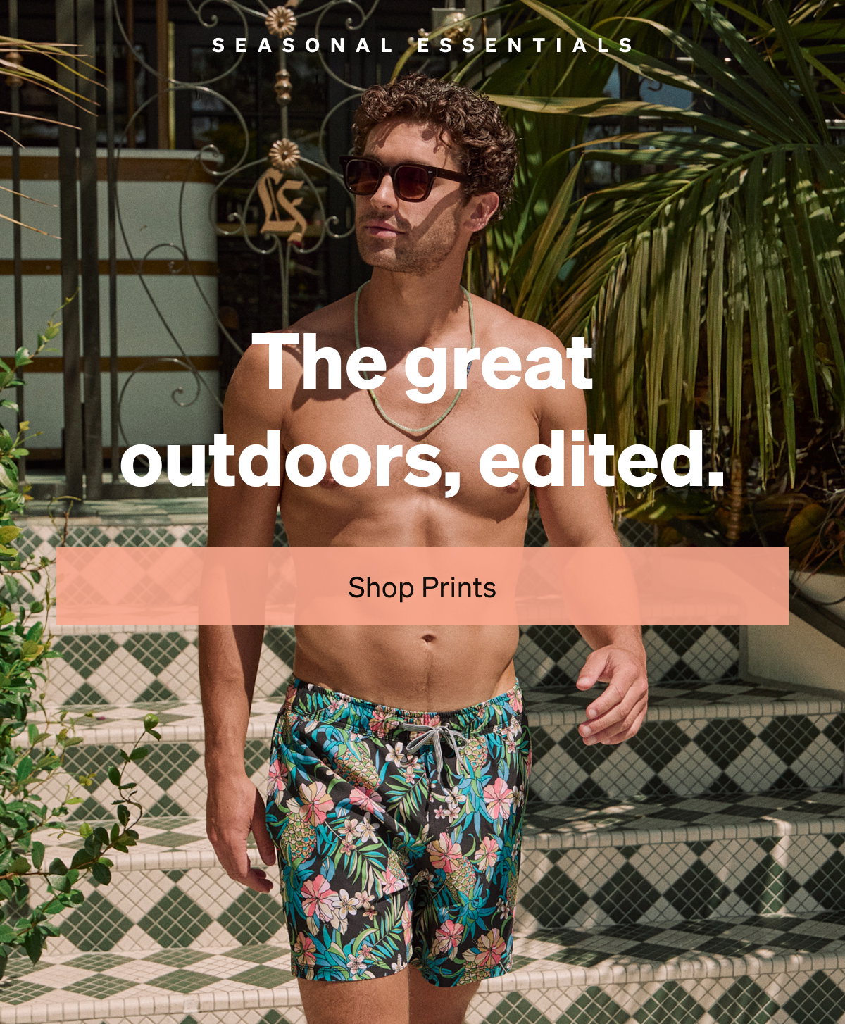 Man wearing floral print swim shorts walks outdoors beside tropical greenery and tiled steps with text reading “The great outdoors, edited.”